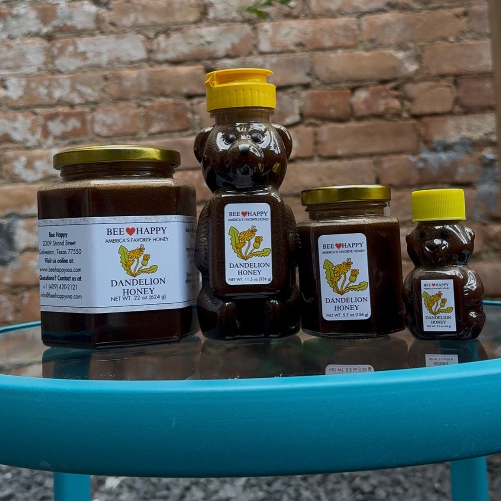 22 oz jar, 11.5 oz bear bottle, 5.5 oz jar and 2.3 oz bear bottle of Bee Happy Dandelion Honey on a glass surface with a brick wall background.