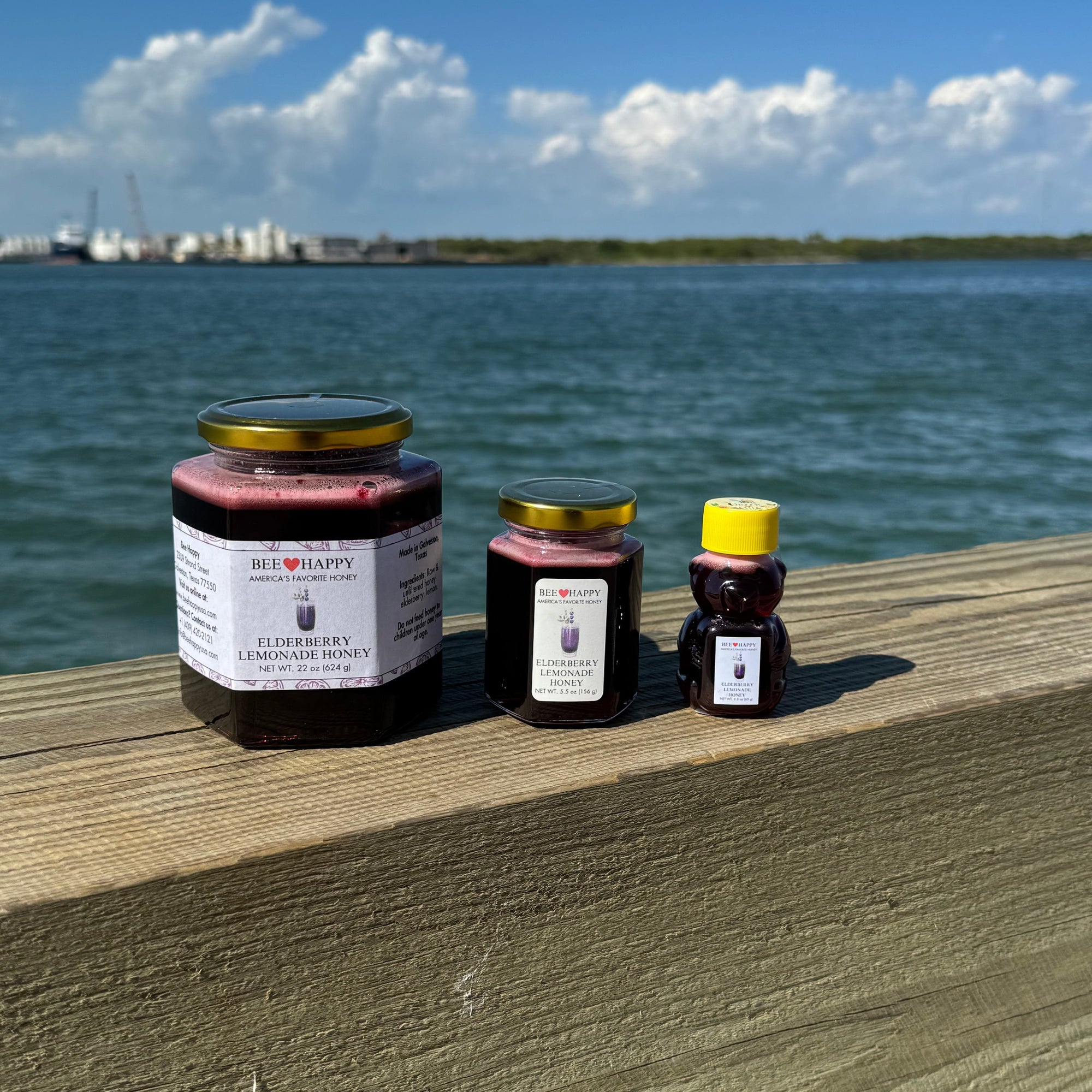 22 oz jar, 5.5 oz jar and 2.3 oz bear bottle of Elderberry Lemonade Honey on a wooden ledge with a waterfront background