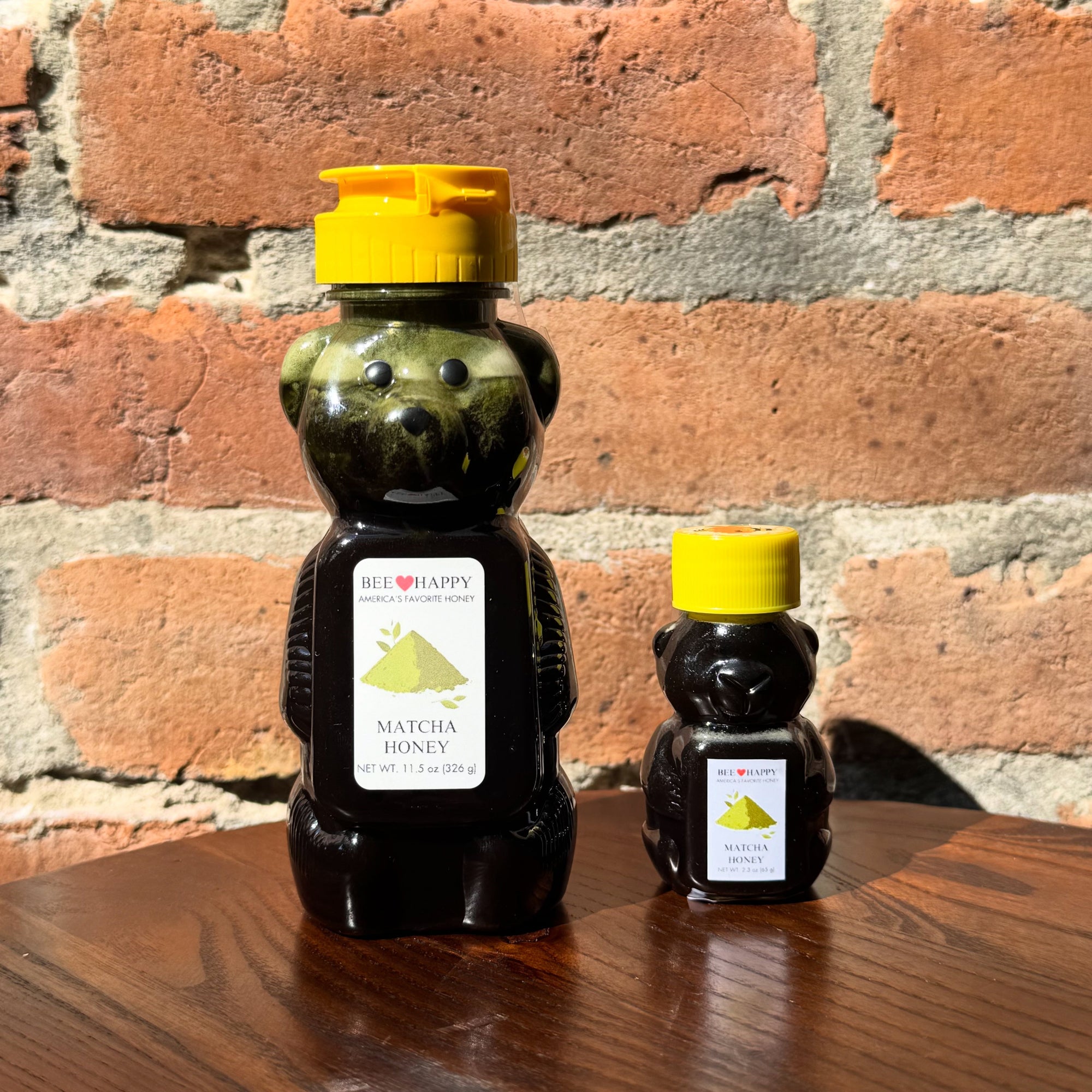 11.5 oz and 2.3 oz bear-shaped honey bottles with yellow caps on a wooden surface against a brick wall.