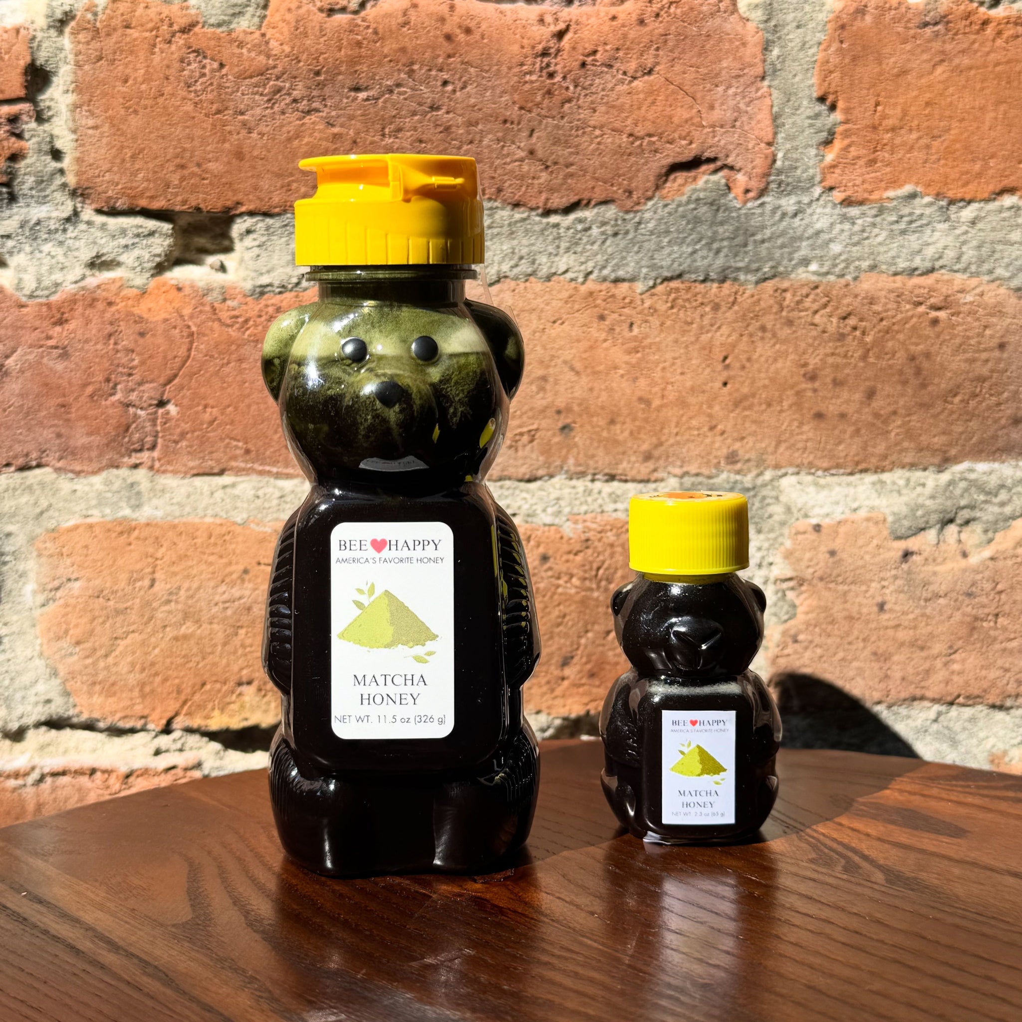 11.5 oz and 2.3 oz bear-shaped honey bottles with yellow caps on a wooden surface against a brick wall.