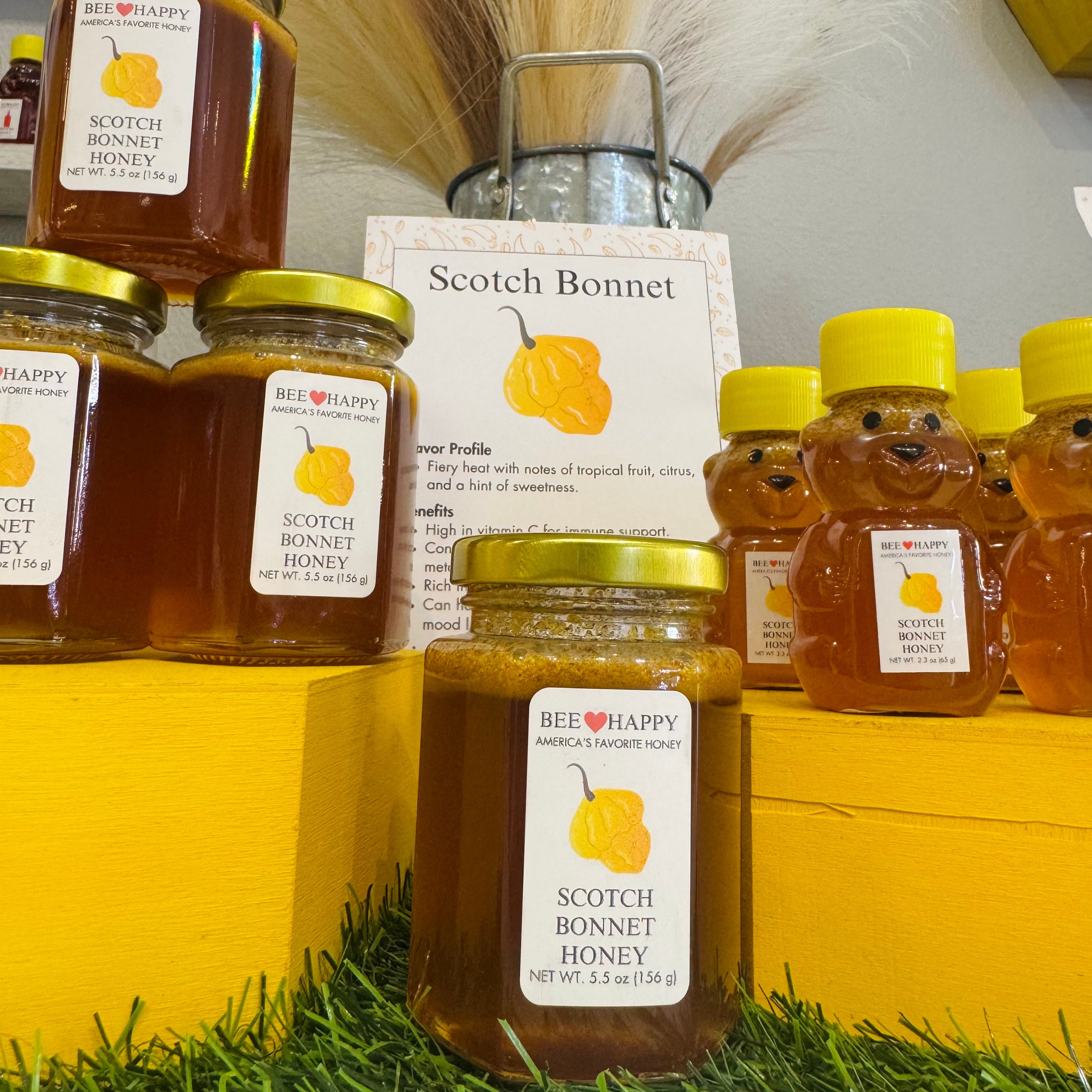 Jars and 2.3 oz bears bottles of Scotch Bonnet Honey on yellow wooden stands.