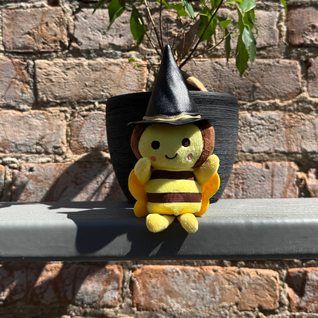 Bee plushie toy with black witch hat sitting on a ledge with a brick wall behind it.