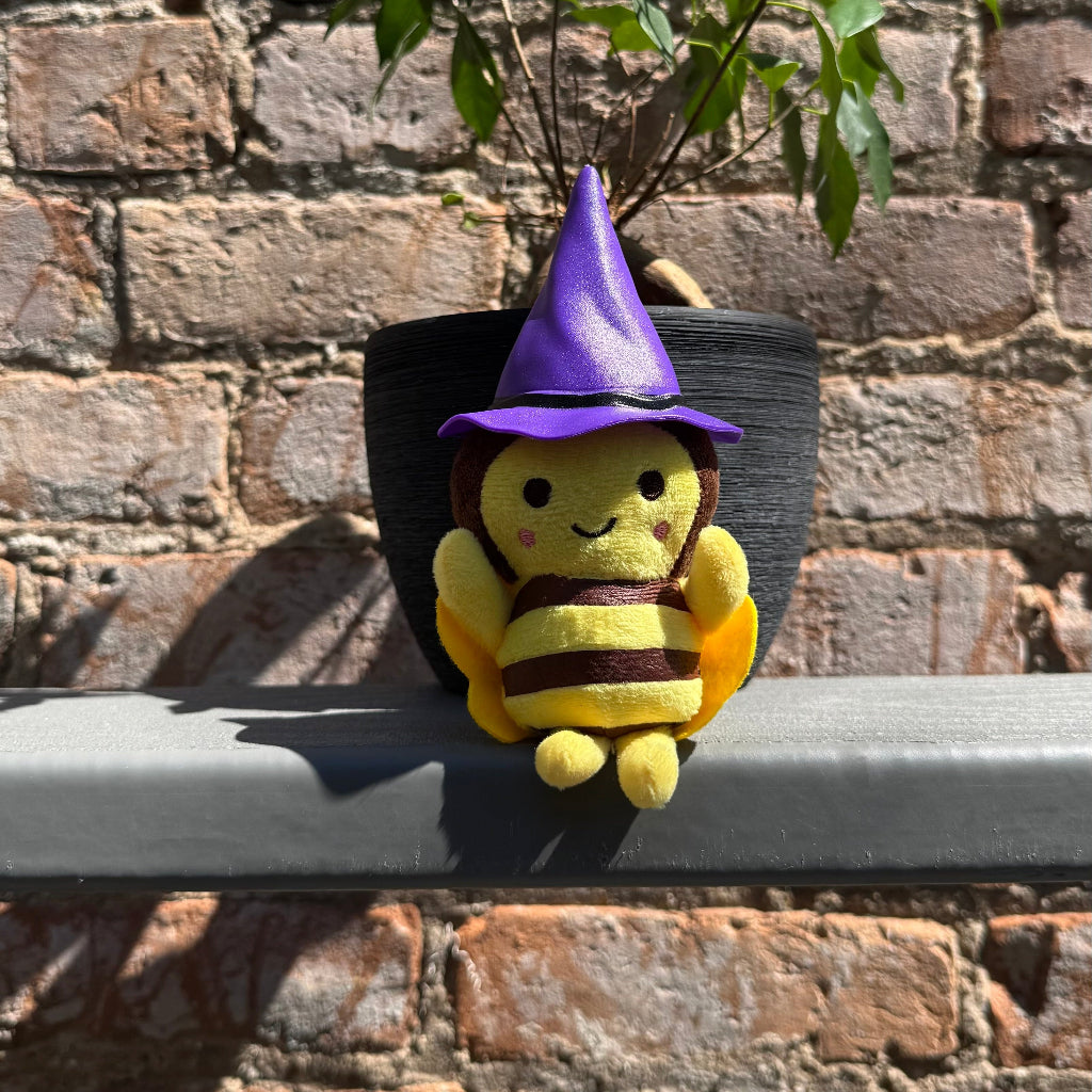 Bee plushie toy with purple witch hat sitting on a ledge with a brick wall behind it.