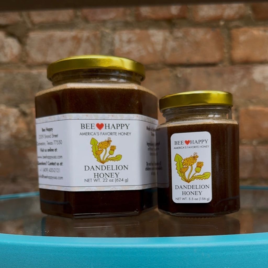 22 oz and 5.5 oz jars of Dandelion Honey from Bee Happy on a glass surface with a brick wall background.