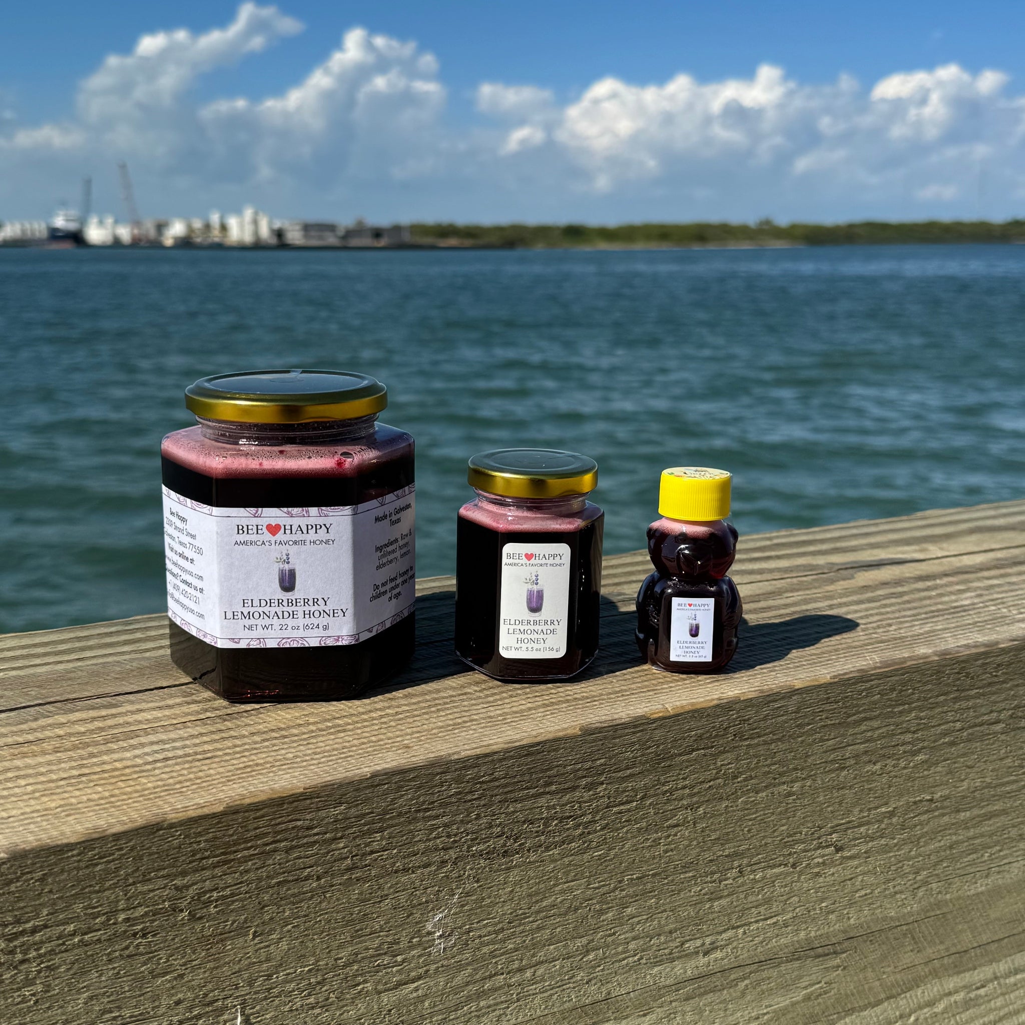 22 oz jar, 5.5 oz jar and 2.3 oz bear bottle of Elderberry Lemonade Honey on a wooden ledge with a waterfront background