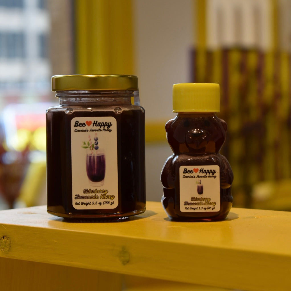 5.5 oz jar and 2.3 oz bear bottle of Elderberry Lemonade Honey on a wooden surface with a blurred background.