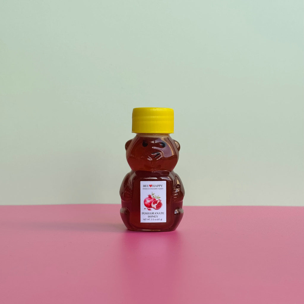 2.3 oz bear bottle of Bee Happy Pomegranate Honey on a pink surface and light green background