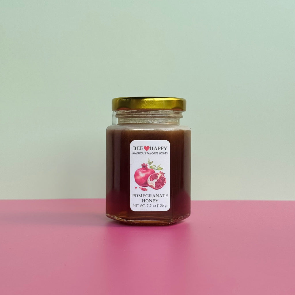 5.5 oz jar of Bee Happy Pomegranate Honey on a pink surface and light green background