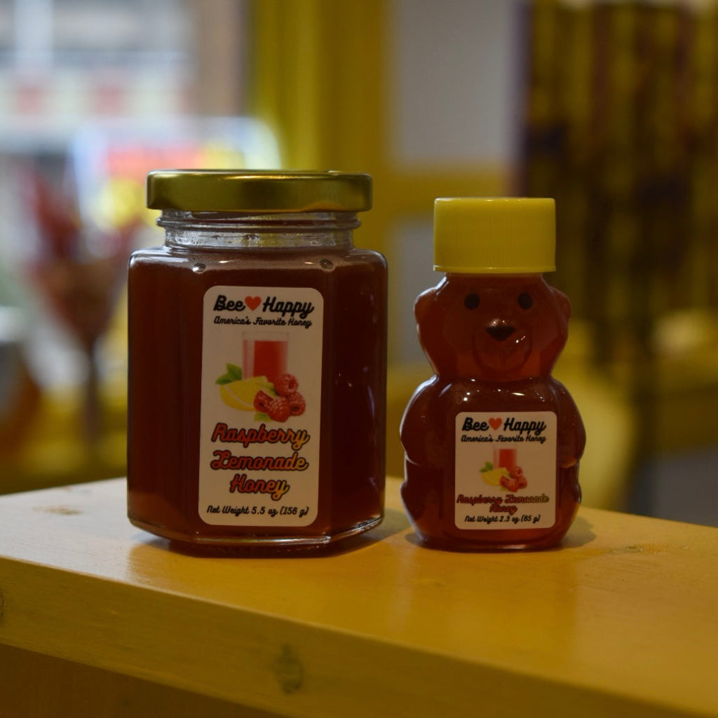 5.5 oz jar and 2.3 oz bear bottle of Raspberry Lemonade Honey on a wooden surface with a blurred background.