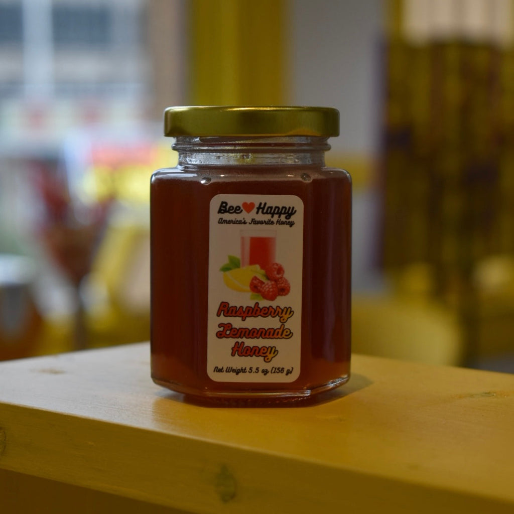 5.5 oz jar of Raspberry Lemonade Honey on a wooden surface with a blurred background.