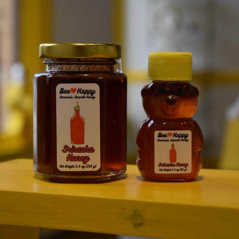 5.5 oz jar and 2.3 oz bear bottle of Sriracha Honey on a wooden surface with a blurred background.