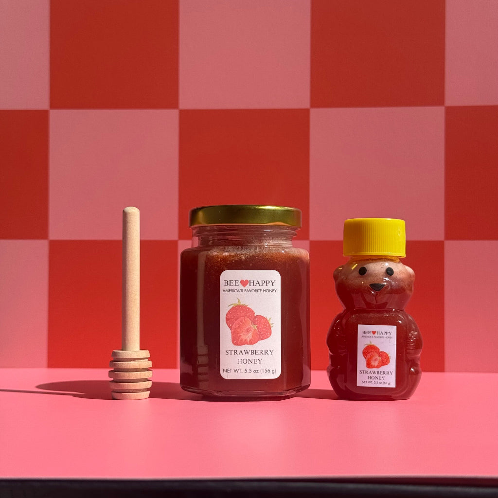 5.5 oz jar and 2.3 oz bear-shaped bottle of Bee Happy Strawberry Honey with a honey dipper on a pink and red checkered background.