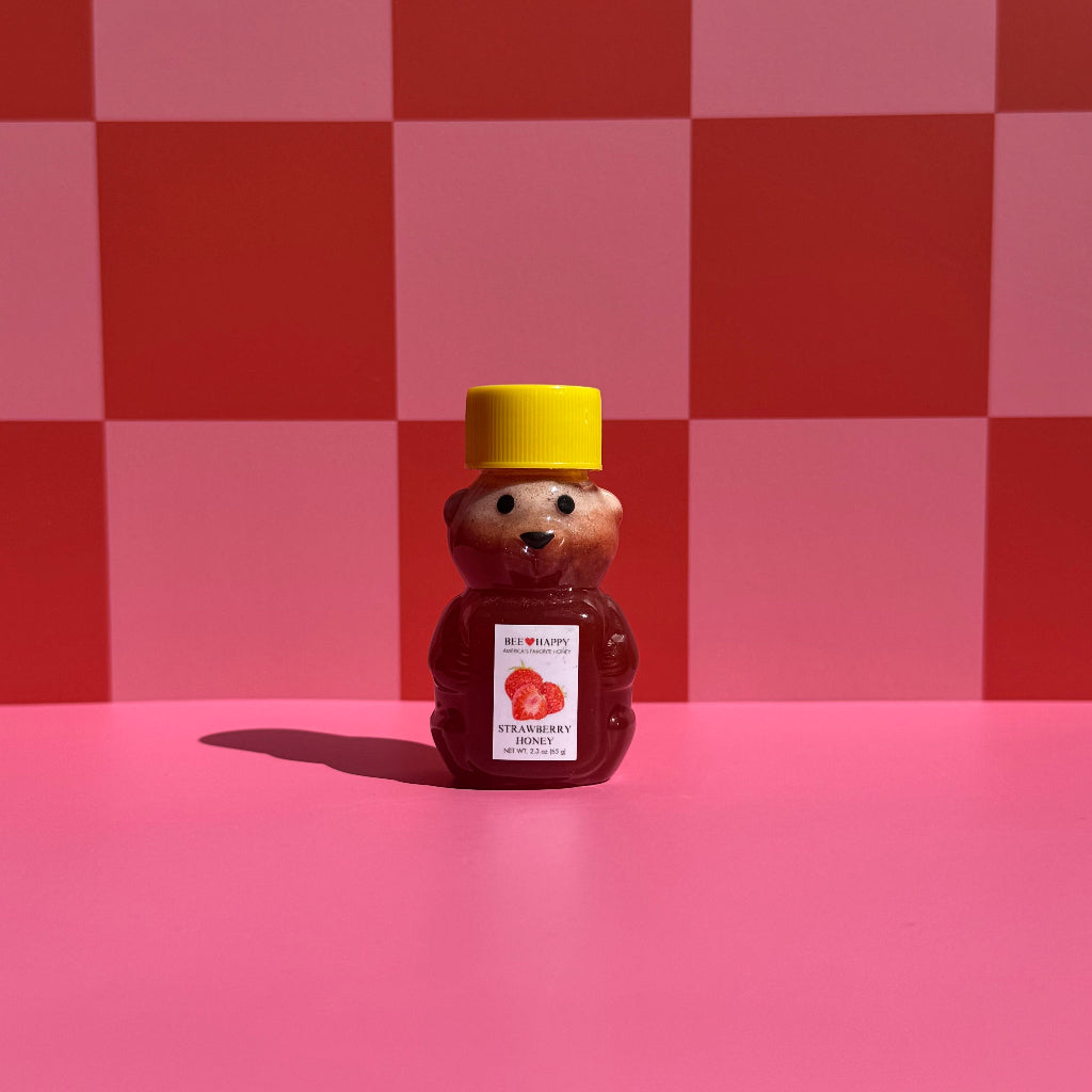 2.3 oz bear-shaped bottle of Bee Happy Strawberry Honey on a pink and red checkered background.