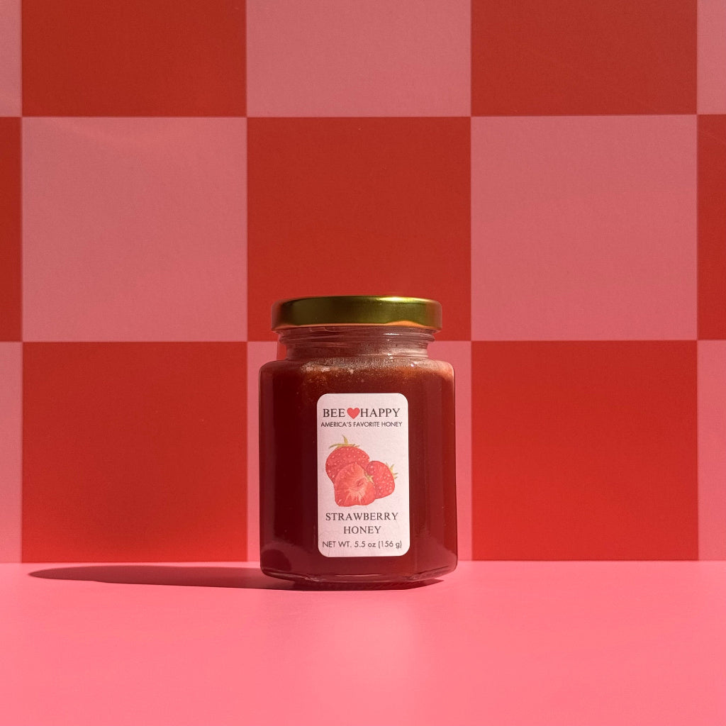 5.5 oz jar of Bee Happy Strawberry Honey on a pink and red checkered background.