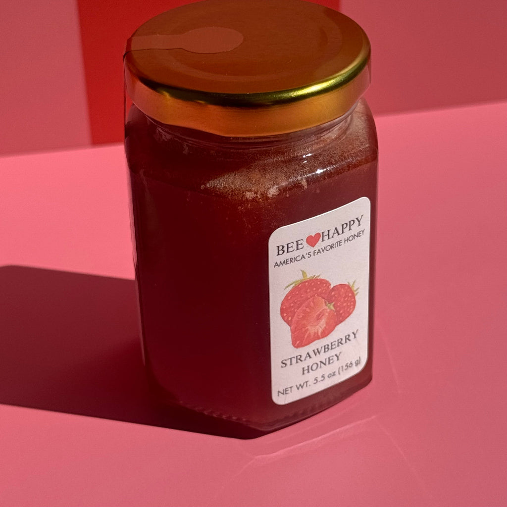 5.5 oz jar of Bee Happy Strawberry Honey on a pink and red checkered background.