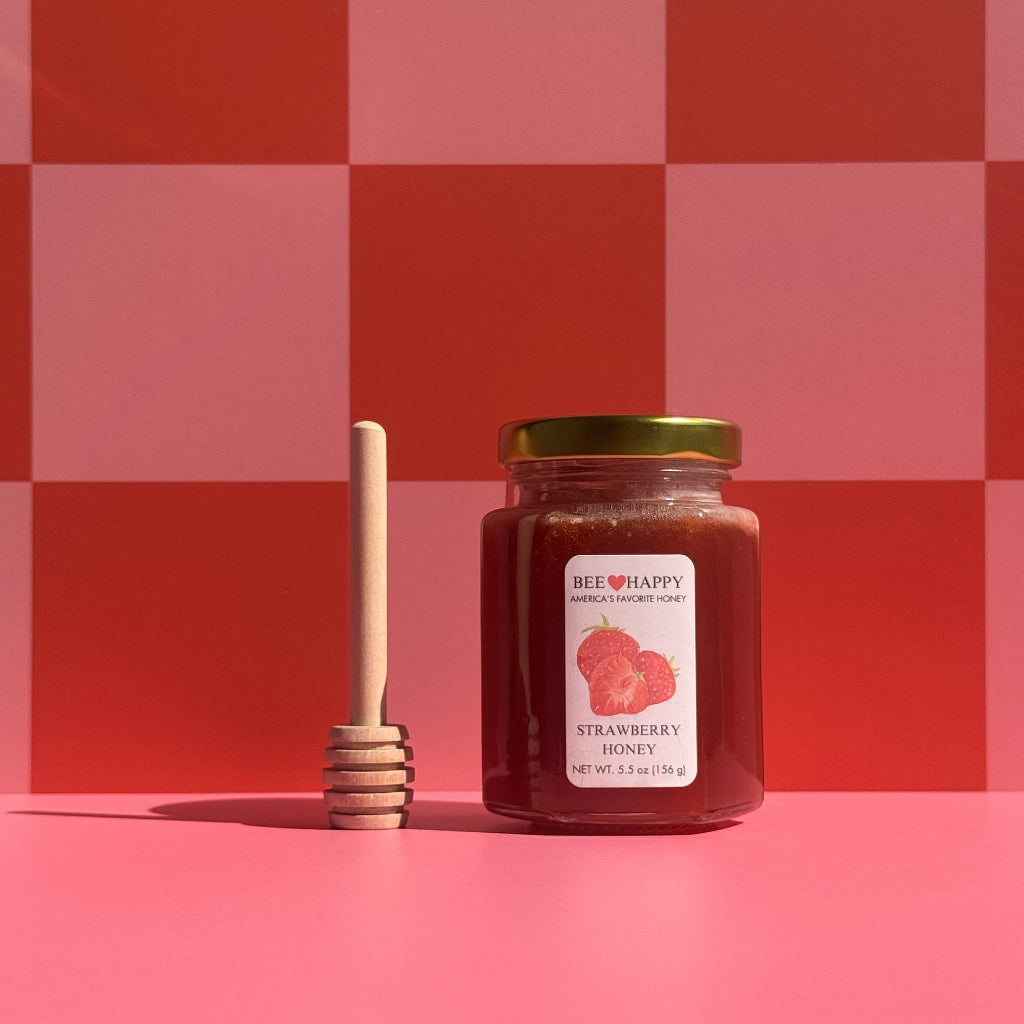 5.5 oz jar of Bee Happy Strawberry Honey with a honey dipper on a pink and red checkered background.