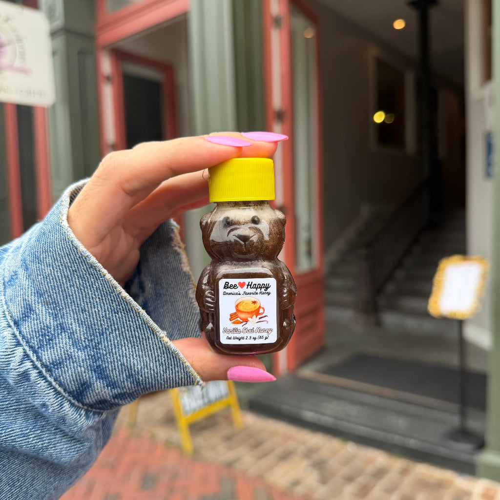 A hand with pink nails holds a 2.3 oz bear bottle of Vanilla Chai Honey in front of a blurred storefront.