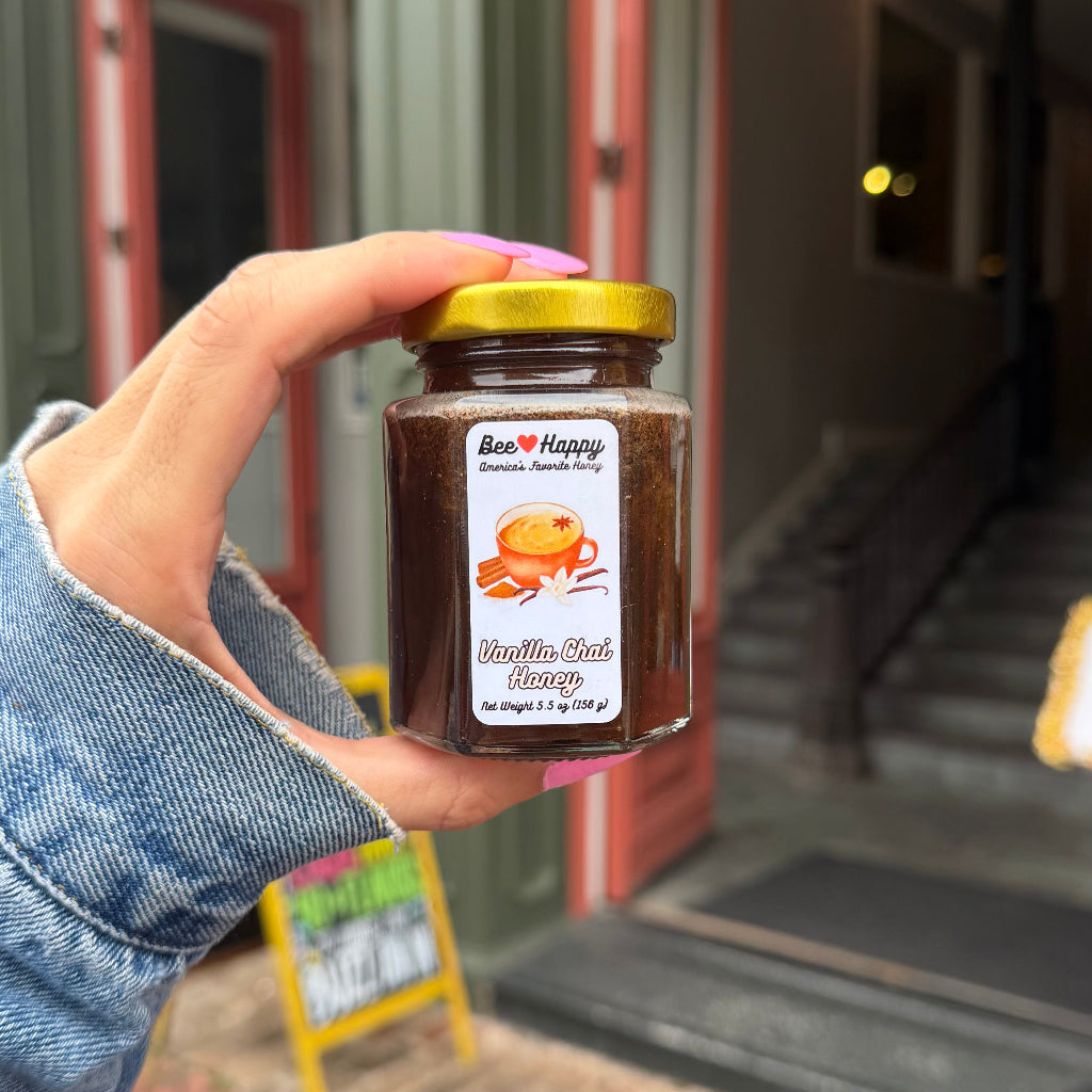 A hand with pink nails holds a 5.5 oz jar of Vanilla Chai Honey in front of a blurred storefront.