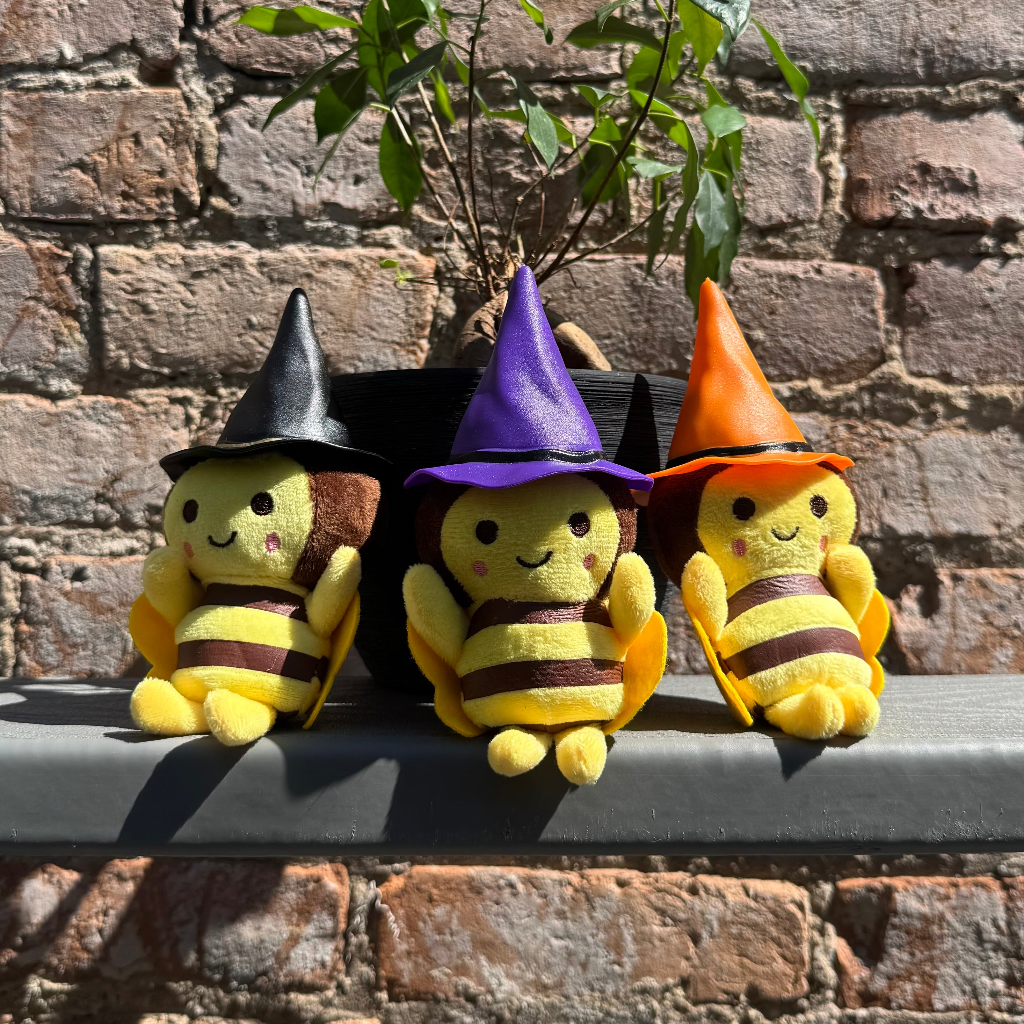 Three plush bee toys with black, purple and orange witch hats sitting on a ledge with a brick wall behind them.