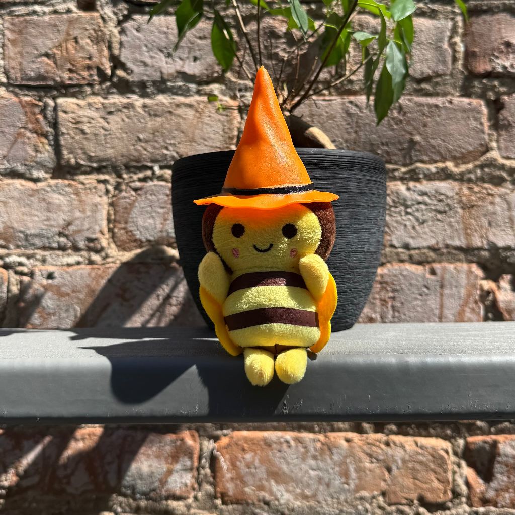 Bee plushie toy with orange witch hat sitting on a ledge with a brick wall behind it.
